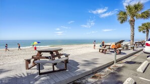 Uma praia com mesas de piquenique e bancos, palmeiras e um céu azul límpido.