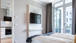 Ein modernes Hotelzimmer mit Bett, Wandfernseher, Küchenzeile und großen Fenstern.