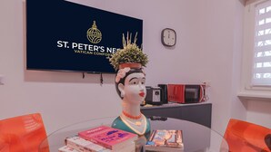 Et værelse med et fjernsyn der viser 'ST. PETER'S NEIGHBORHOOD VATICAN COMFORT', en potteplante på et bord, bøger, en mikrobølgeovn og et ur på væggen.