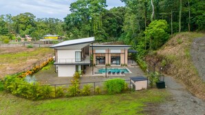 Ein modernes, zweistöckiges Haus mit Swimmingpool, umgeben von üppiger Vegetation und einer hügeligen Landschaft.