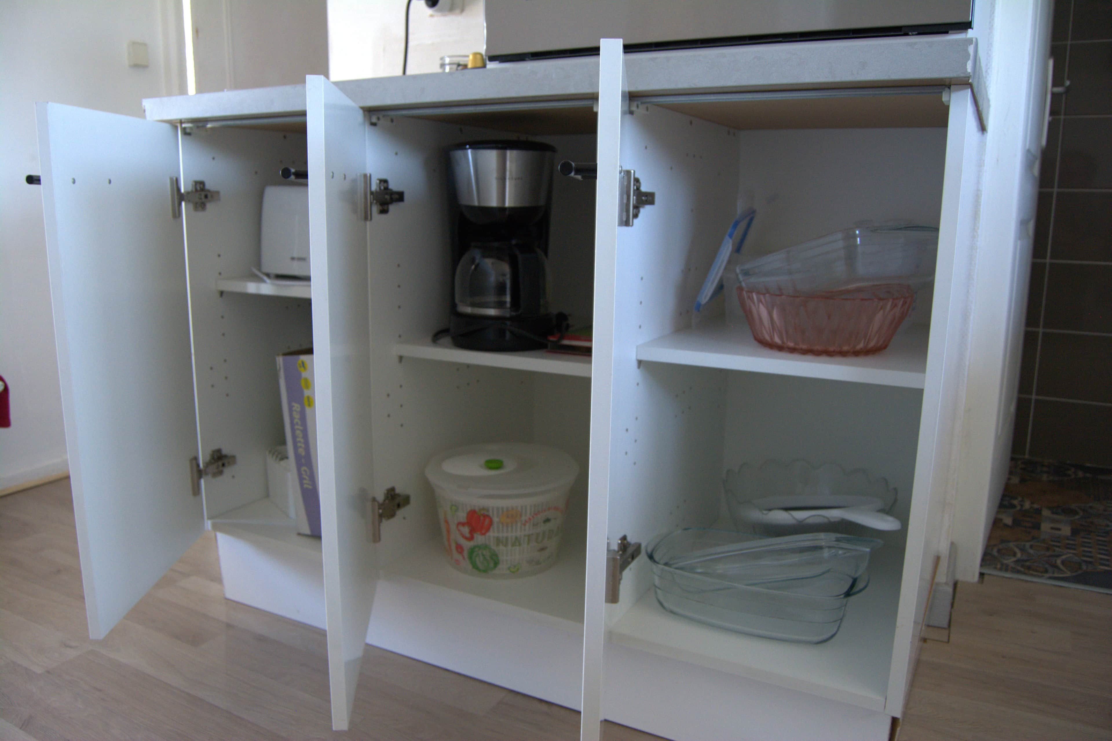 A white kitchen cabinet with open doors, revealing a coffee maker, a container with a lid, and a clear plastic dish rack.