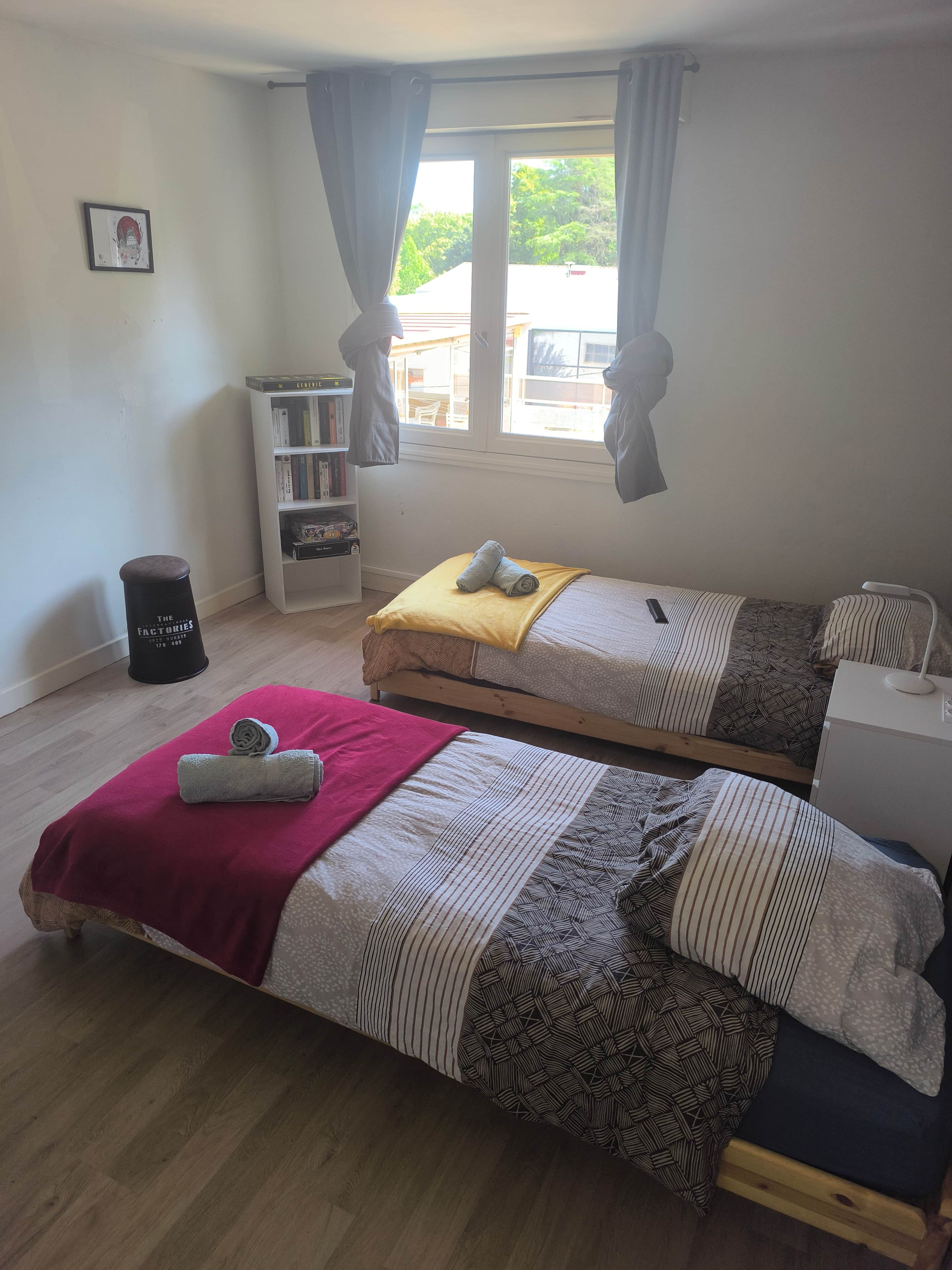 A bedroom with two single beds, a bookshelf, a waste bin, and a window with curtains.