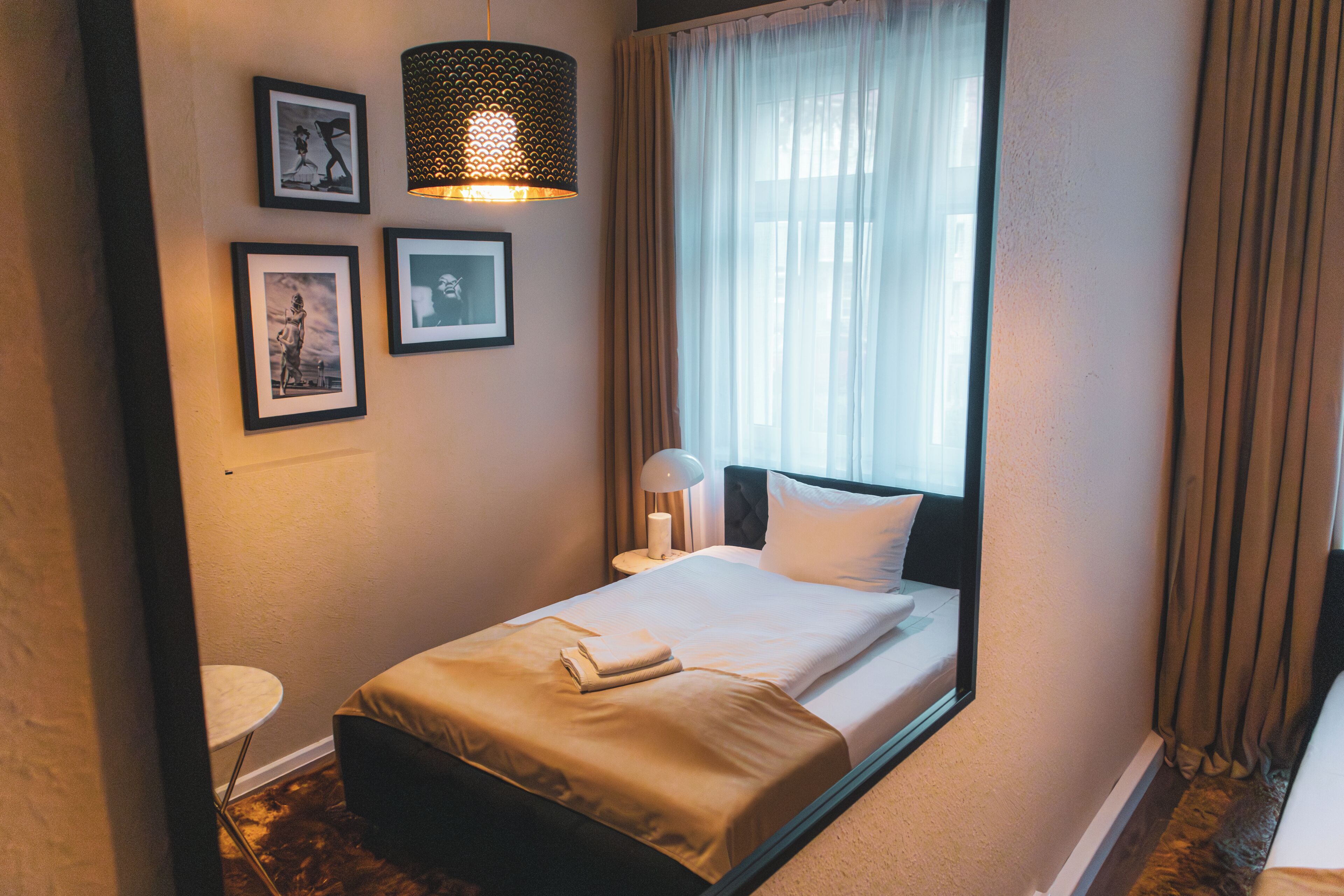 A neatly made bed with a white comforter and a folded towel, a bedside lamp, and a wall-mounted picture frame.