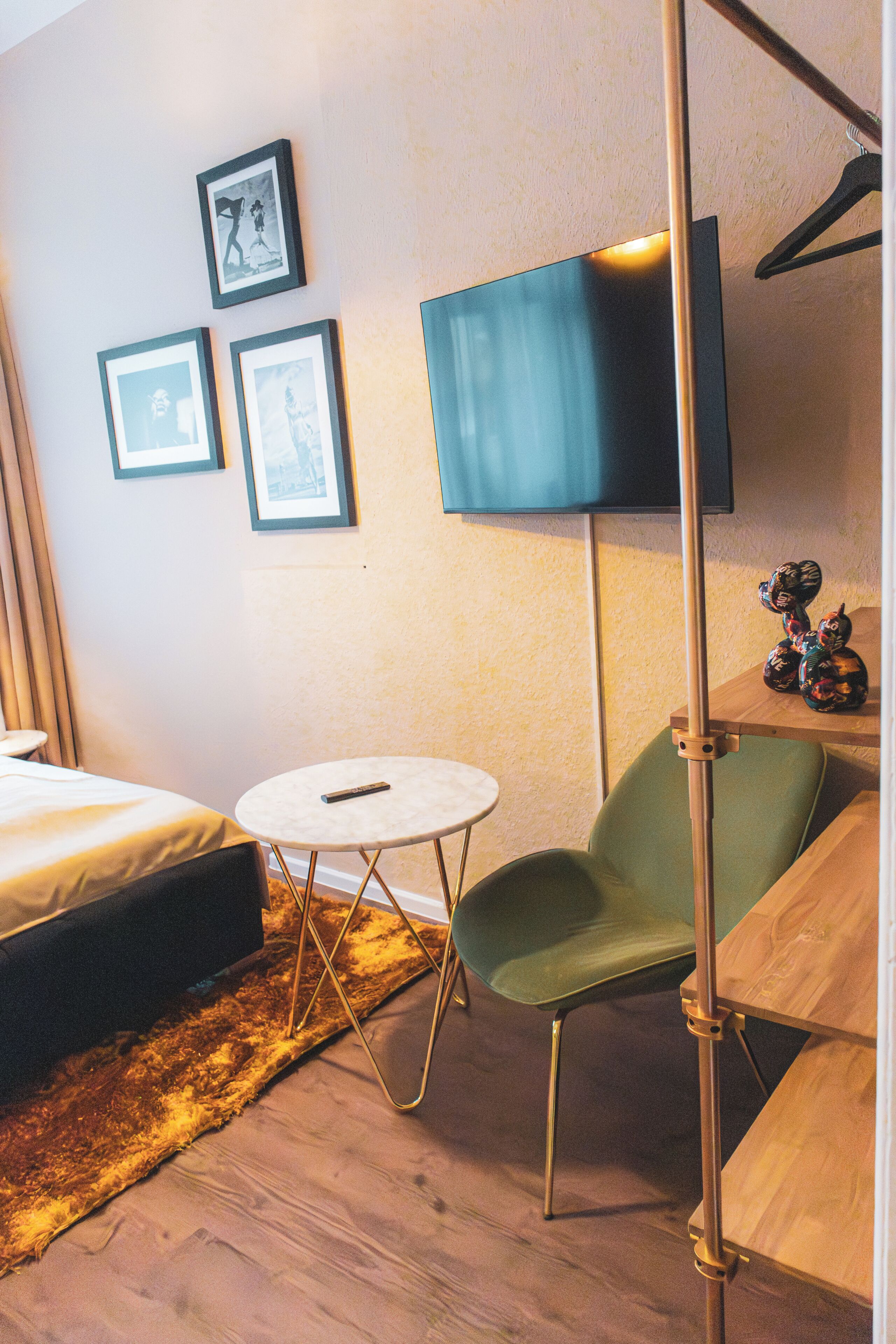 A hotel room with a bed, a small round table, a green chair, a floor lamp, and a flat-screen TV mounted on the wall.