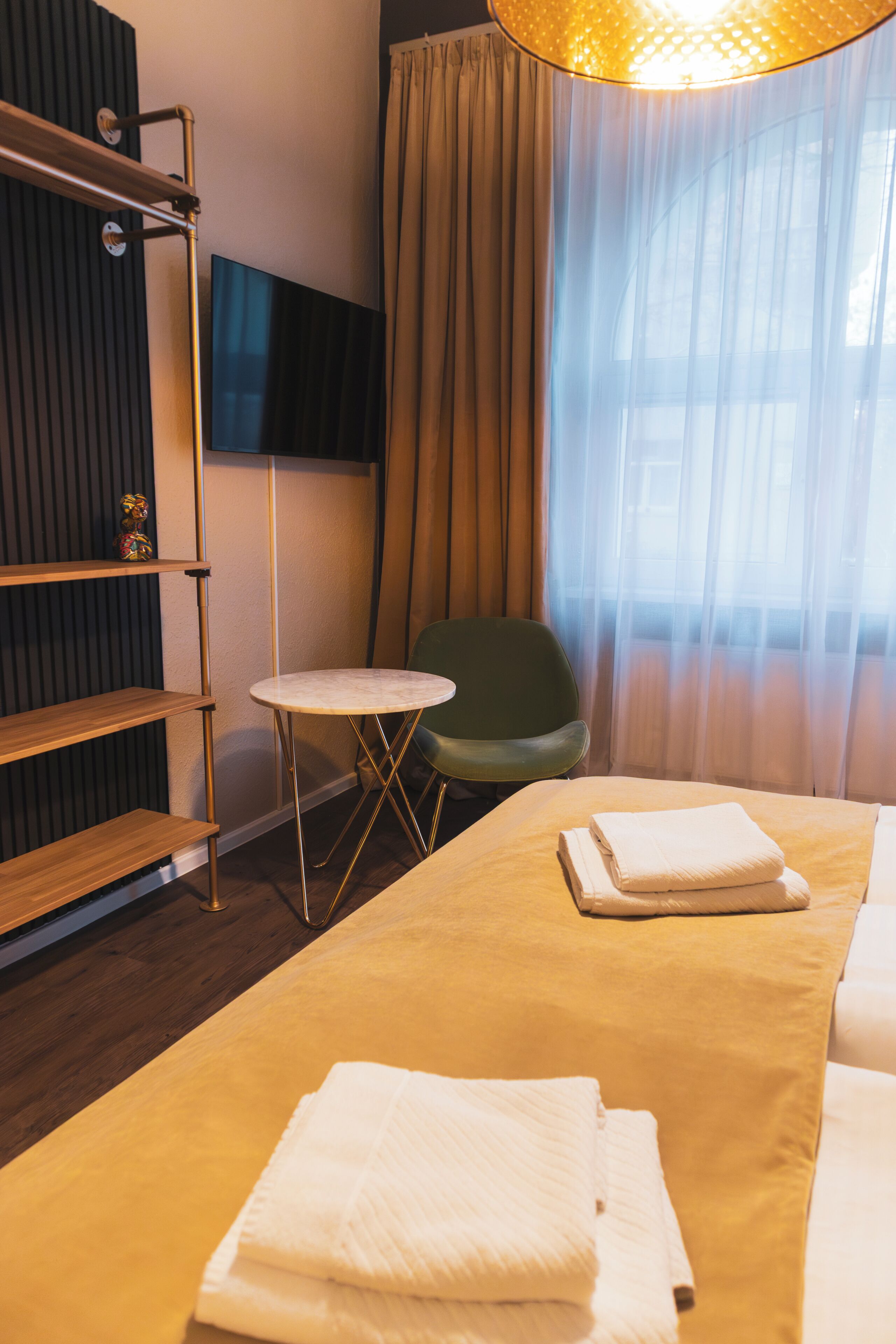 A hotel room with a bed, a small round table, a chair, a TV mounted on the wall, and a window with curtains.