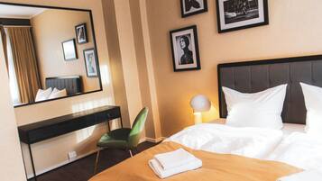 A hotel room with a bed, a desk, a chair, a lamp, and framed pictures on the wall.