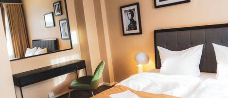 A hotel room with a bed, a desk, a chair, a lamp, and framed pictures on the wall.