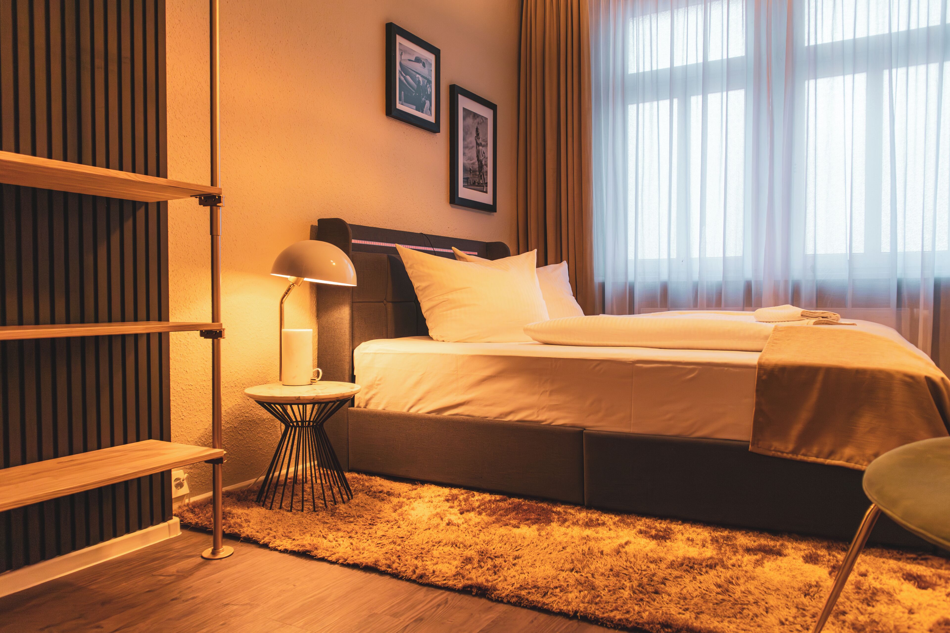 Habitación de hotel con una cama, una mesita de noche con lámpara, una silla, una pequeña mesa y un ventanal con cortinas.