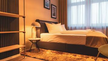 Habitación de hotel con una cama, una mesita de noche con lámpara, una silla, una pequeña mesa y un ventanal con cortinas.