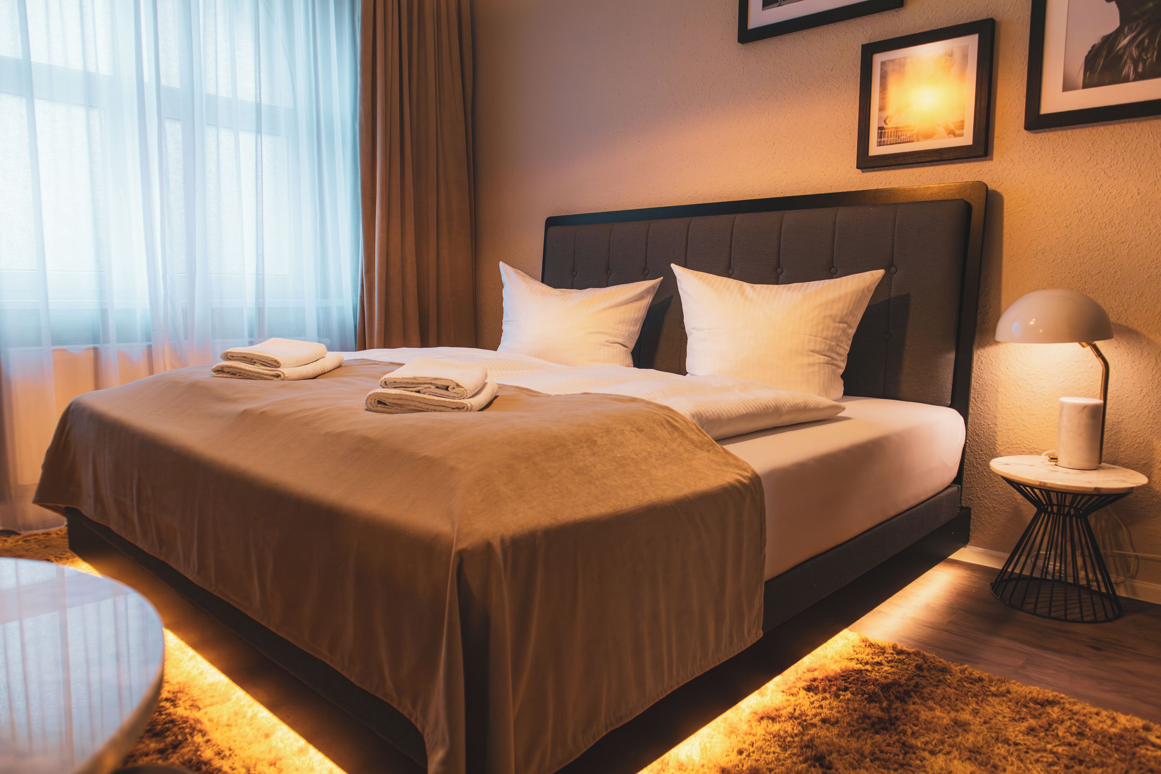 A neatly made bed with pillows and a blanket, a bedside table with a lamp, and a window with sheer curtains.