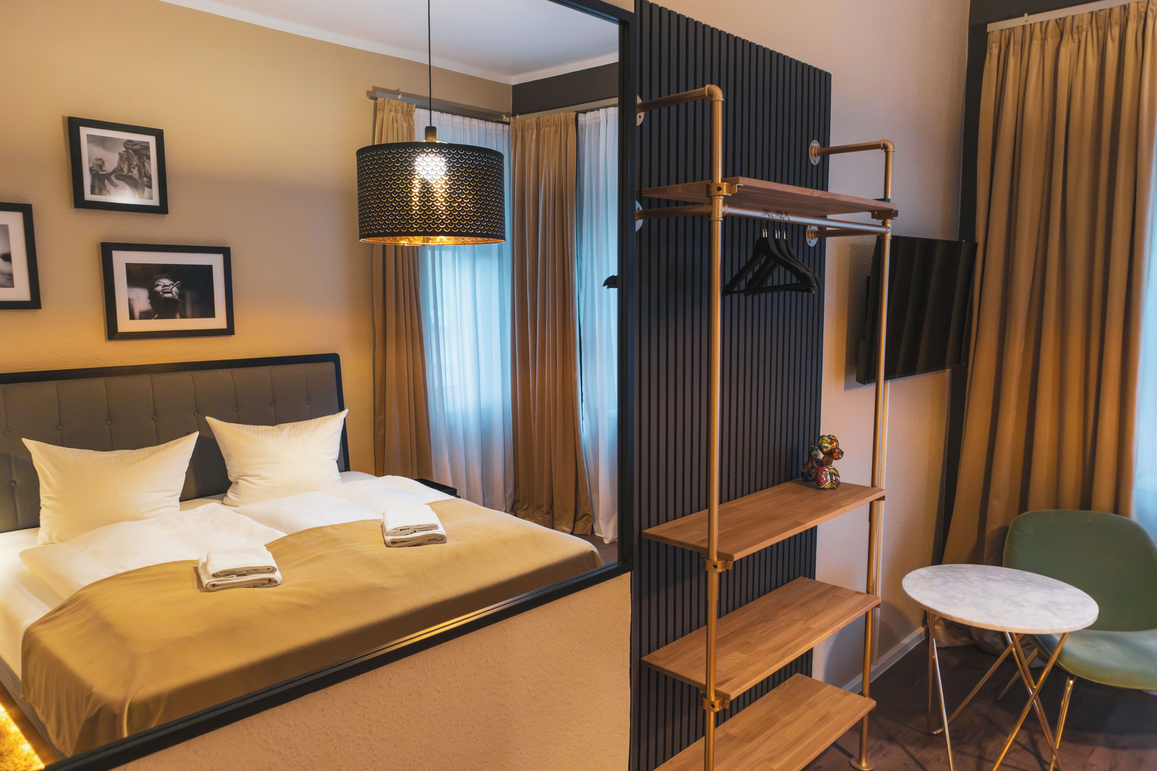 A hotel room with a bed, a hanging light fixture, a TV mounted on the wall, and a small table with a chair.