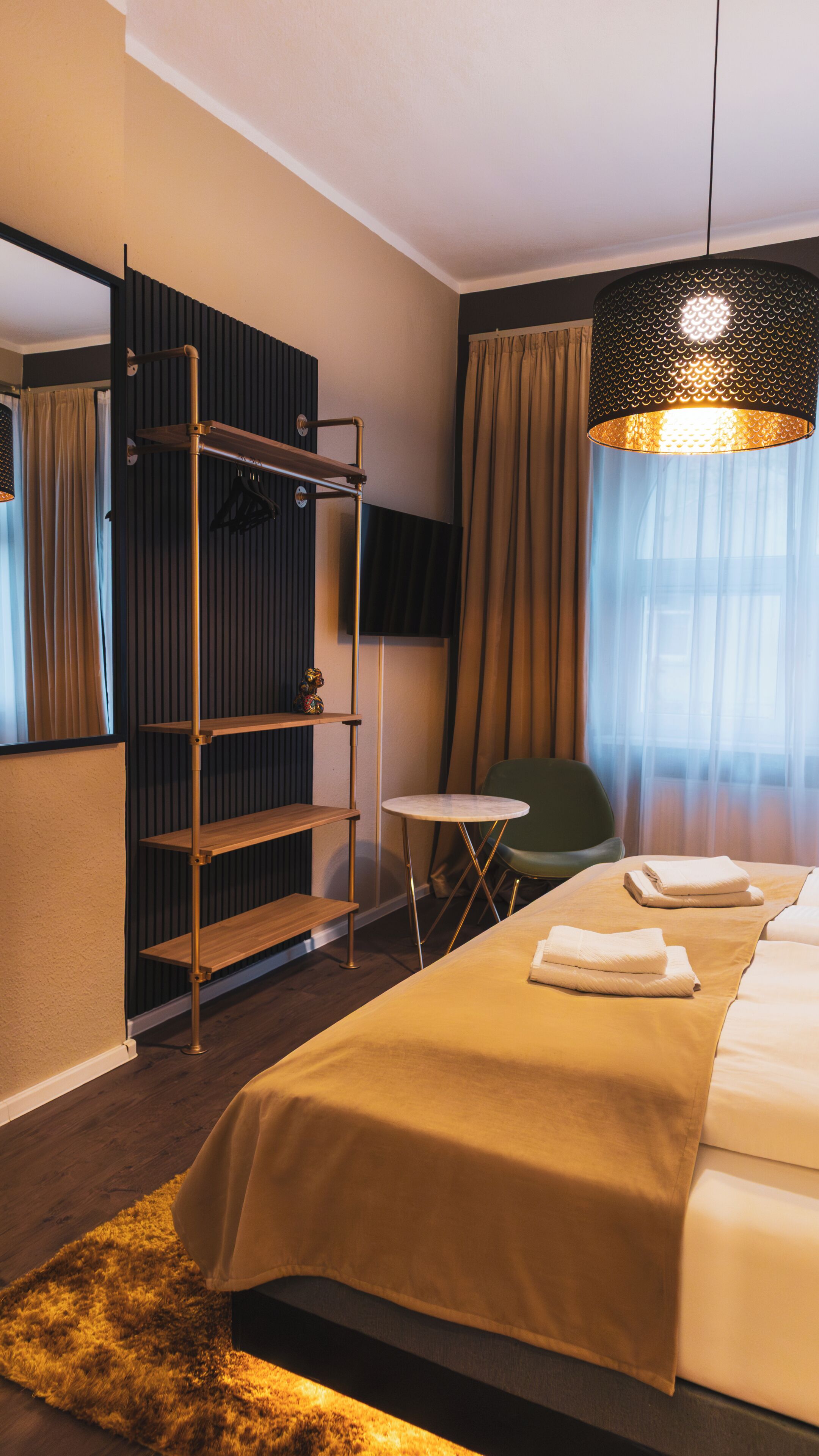 A hotel room with a bed, a small table, a chair, a shelf unit, and a hanging light fixture.