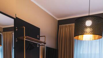 A hotel room with a bed, a small table, a chair, a shelf unit, and a hanging light fixture.