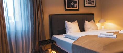 Una habitación de hotel con cama, mesita de noche, lámpara y cortinas.