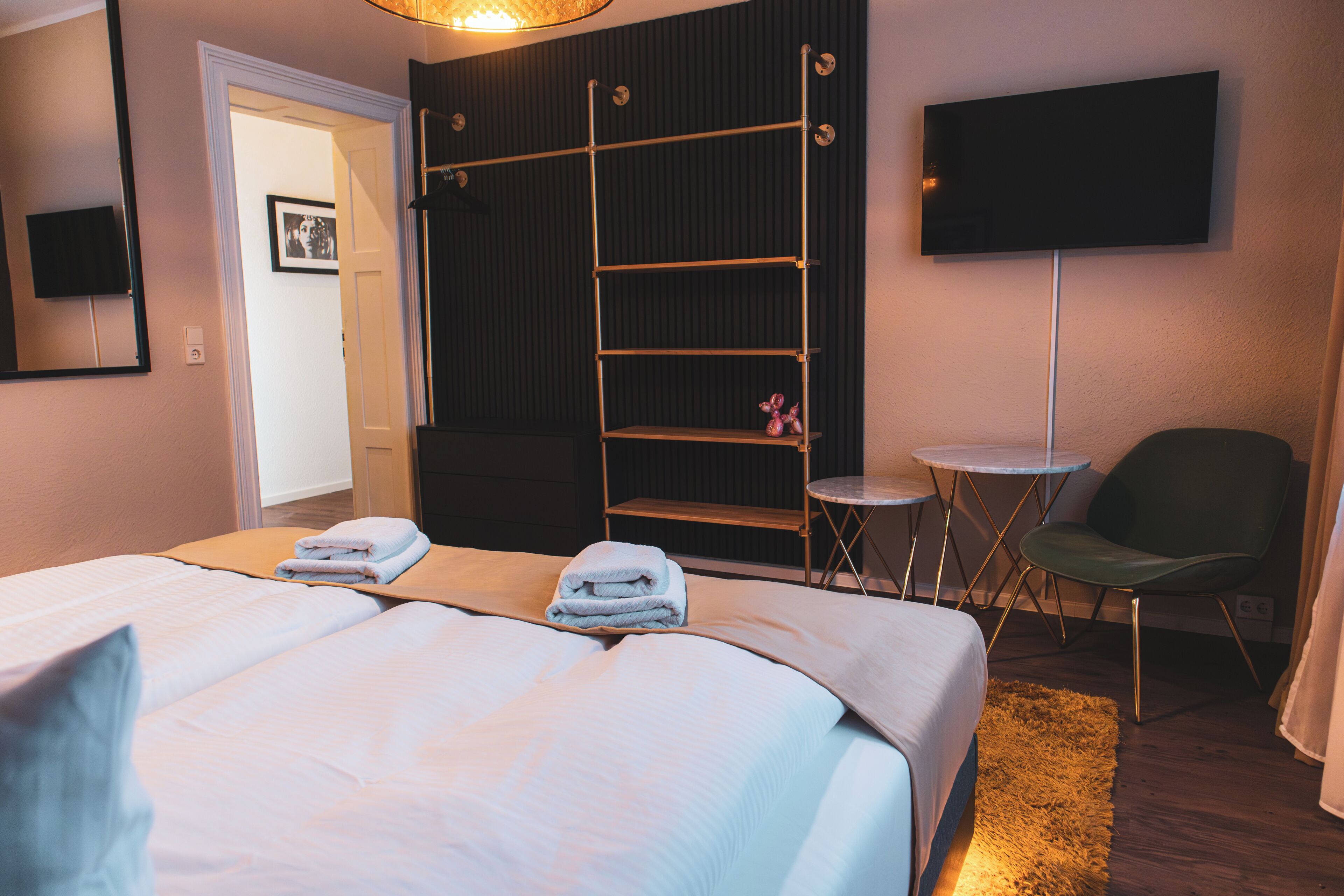 Ein Hotelzimmer mit einem Bett, einem an der Wand montierten Fernseher, einem Sessel, einem kleinen Tisch und einem Regal mit Handtüchern.