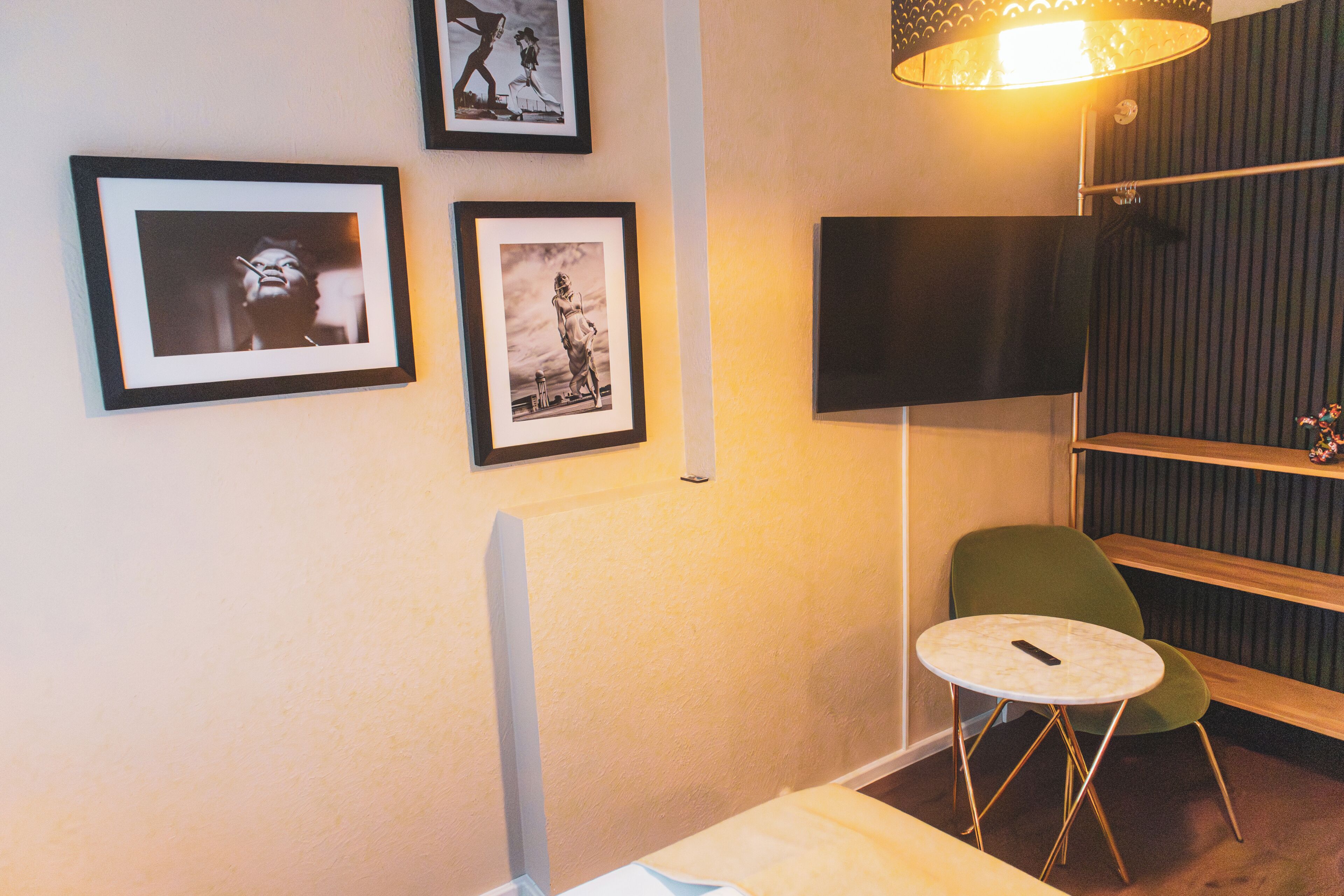 Una habitación con una cama, una mesita redonda pequeña, una silla verde, un televisor de pantalla plana montado en la pared y dos cuadros enmarcados.