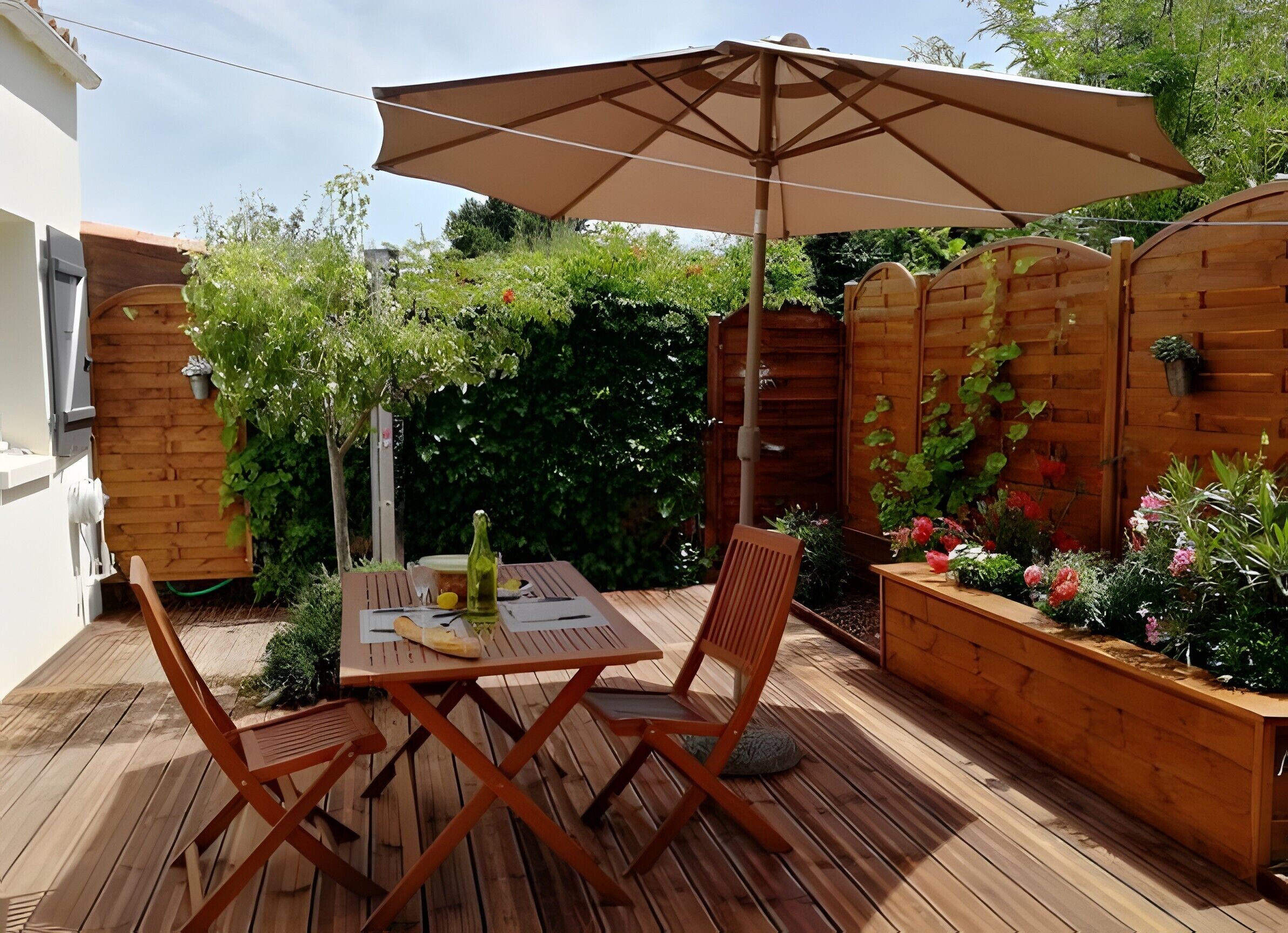 A wooden deck with a table and chairs, an umbrella, and a wooden fence with plants.