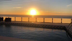 A pool with a view of the sunset over the ocean, a tiled deck, and a railing.