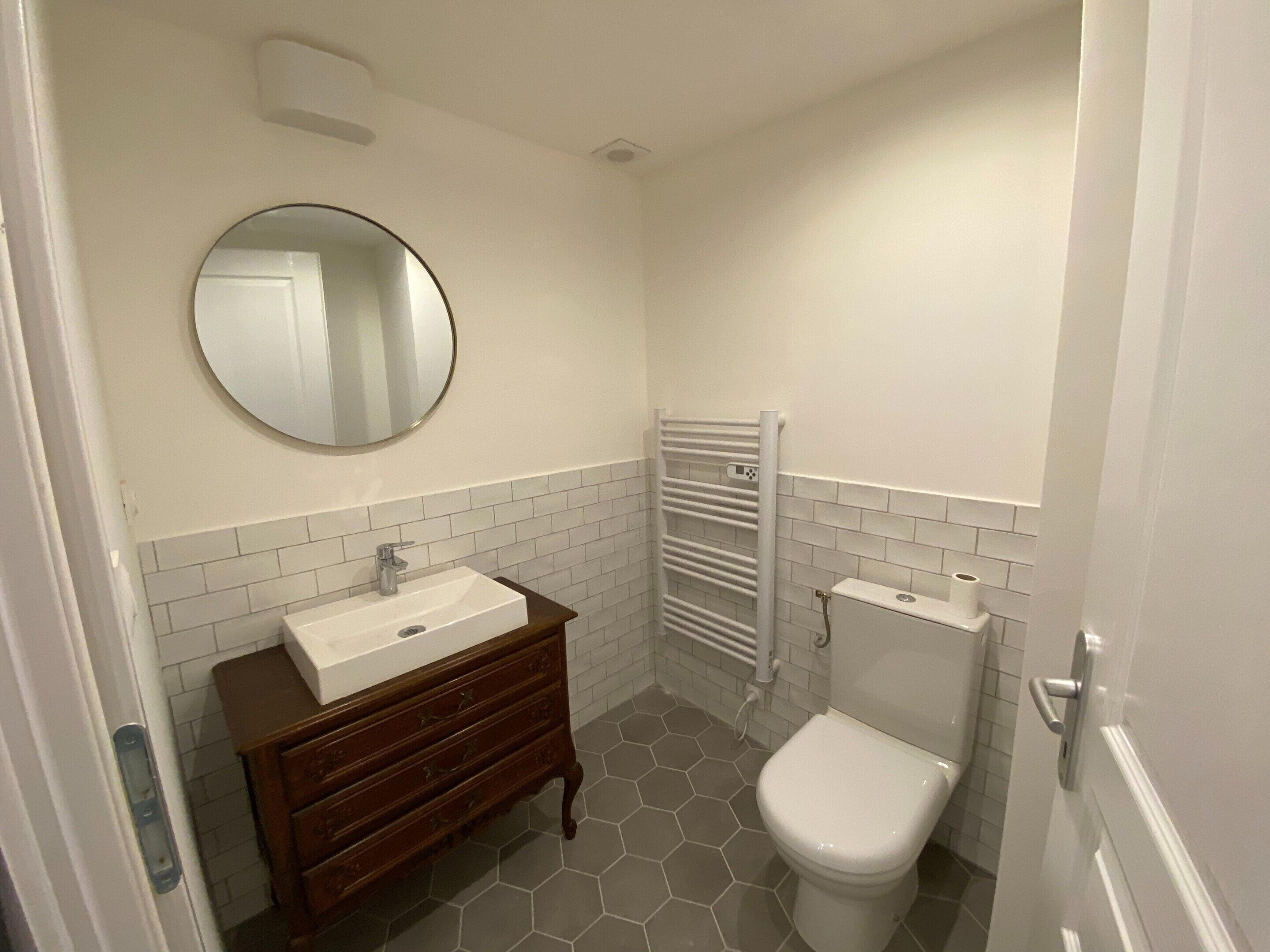 Un baño con lavamanos blanco, tocador de madera, espejo redondo, inodoro blanco y un perchero para toallas.