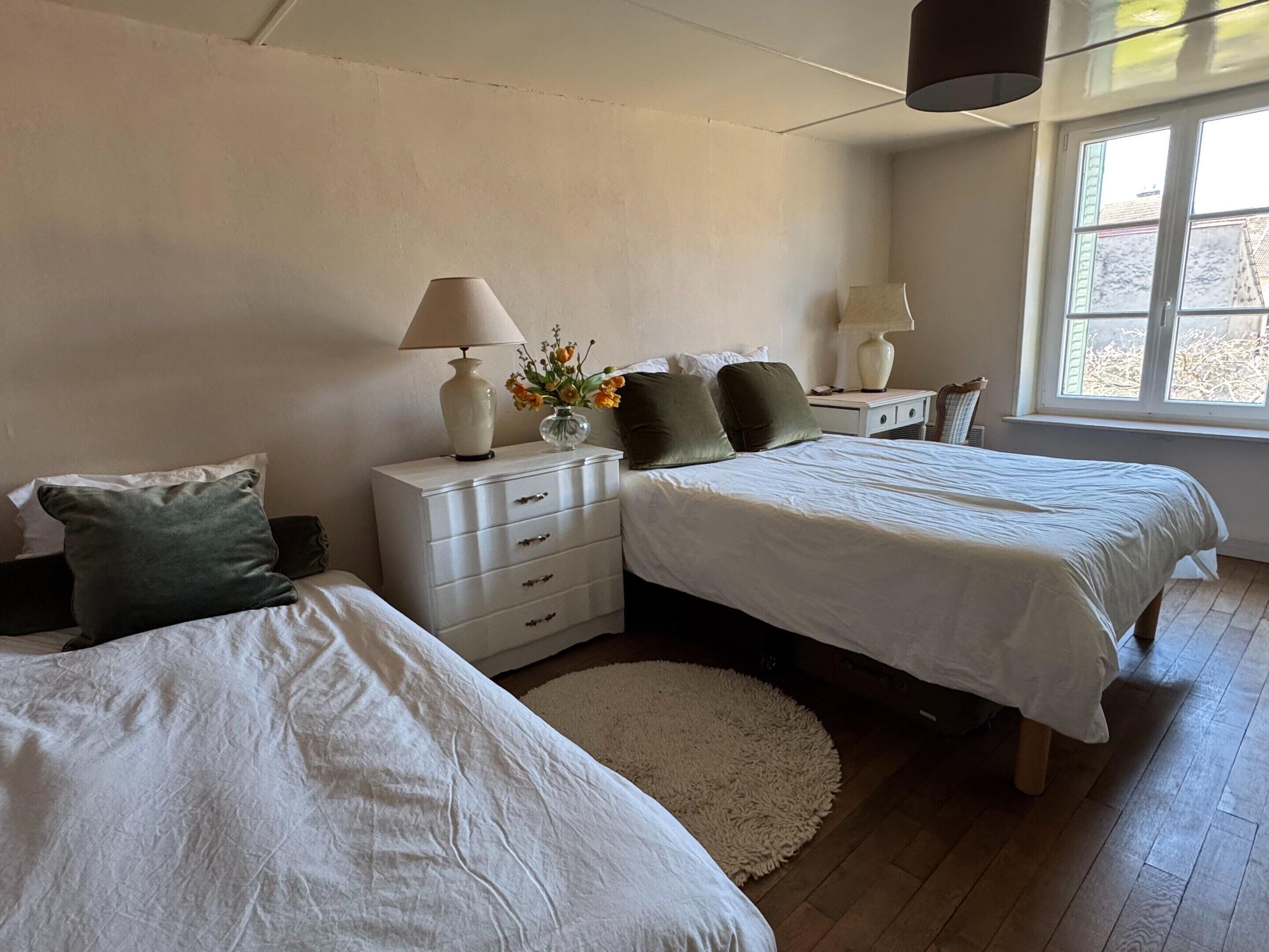A bedroom with two beds, a nightstand with lamps, a vase with flowers, and a window with a view.