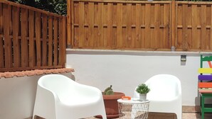 Eine Terrasse mit zwei weißen Plastikstühlen, einem kleinen runden Tisch mit einem Blumentopf und einem bunten Stuhl.