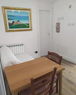 Una habitación con cama, una mesa y una silla. Hay un cuadro en la pared, un radiador y una puerta.