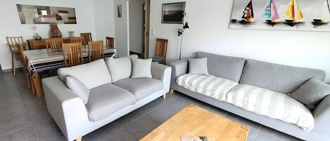 A modern living room with a grey sofa, wooden dining table, and chairs, a coffee table with a magazine, and a painting of sailboats on the wall.
