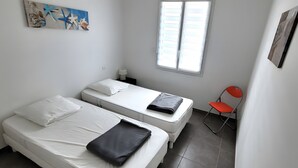 Une petite pièce propre, avec deux lits, une fenêtre, une chaise et un tableau accroché au mur.