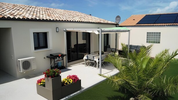 Une maison moderne avec un toit en tuiles, un patio couvert avec des meubles d’extérieur et un jardin agrémenté de jardinières.