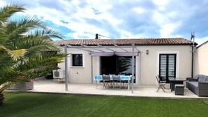 Une maison dotée d’un patio couvert, d’une table à manger, de chaises, d’un canapé et d’un petit jardin agrémenté de palmiers.