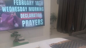 A hotel room with a bed, a flat-screen TV displaying a message, a plant, and a window with curtains. - Executive Studio Room at Bantree Apartments,Accra Airport City (Accra)