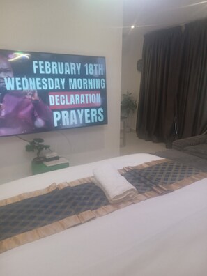 A hotel room with a bed, a flat-screen TV displaying a message, a plant, and a window with curtains. - Executive Studio Room at Bantree Apartments,Accra Airport City (Accra)
