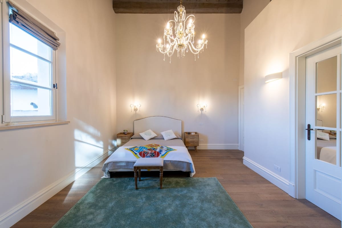 Ein Schlafzimmer mit einem Bett, einer Chandelieber, Nachttischlampen, einer Bank, einem Fenster und einer Tür.