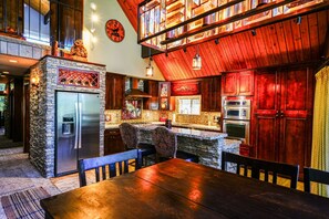 A kitchen with stone countertops, wooden cabinets, a stainless steel refrigerator, and a dining area with a wooden table and chairs. - Wolf Lodge in the Pines (Running Springs)