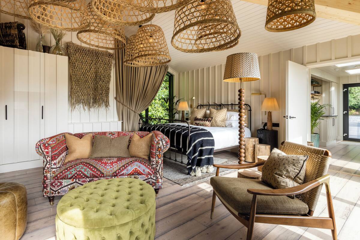 Amplia sala de estar con sofá, reposapiés y sillones, cama con ropa de cama estampada y un ventanal con cortinas.
