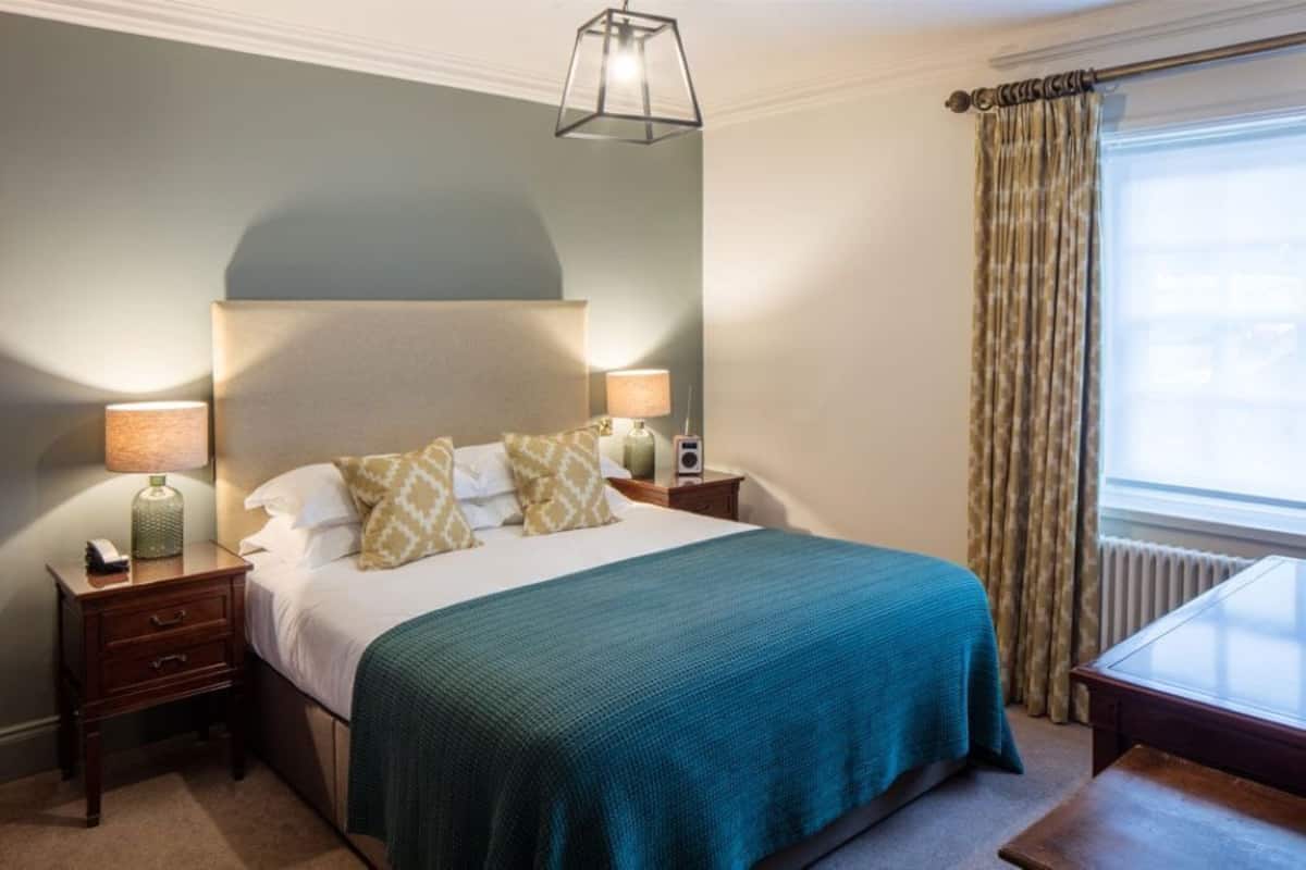 Una habitación de hotel con una cama, lámparas de noche, una mesa y una ventana con cortinas.