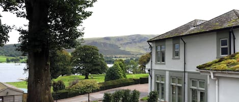 A building with a white exterior, a large tree in the foreground, and a view of a lake and hills in the background.