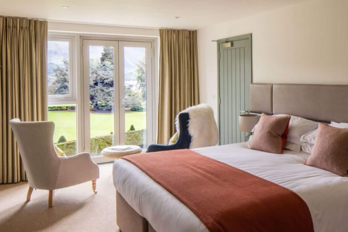 Habitación de hotel con una cama grande, una silla, una mesita y vistas a un jardín a través de una puerta de vidrio.