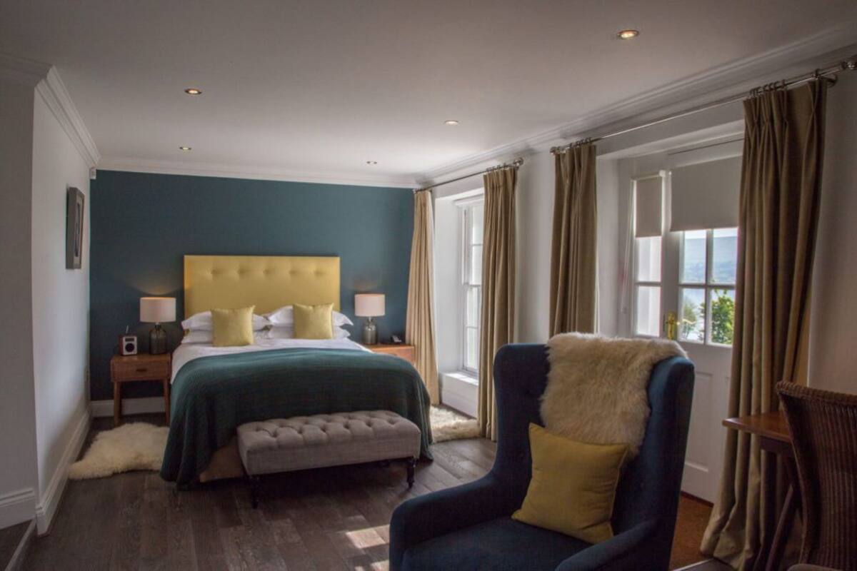 Un dormitorio con una cama grande, mesitas de noche, una silla y un ventanal con cortinas.