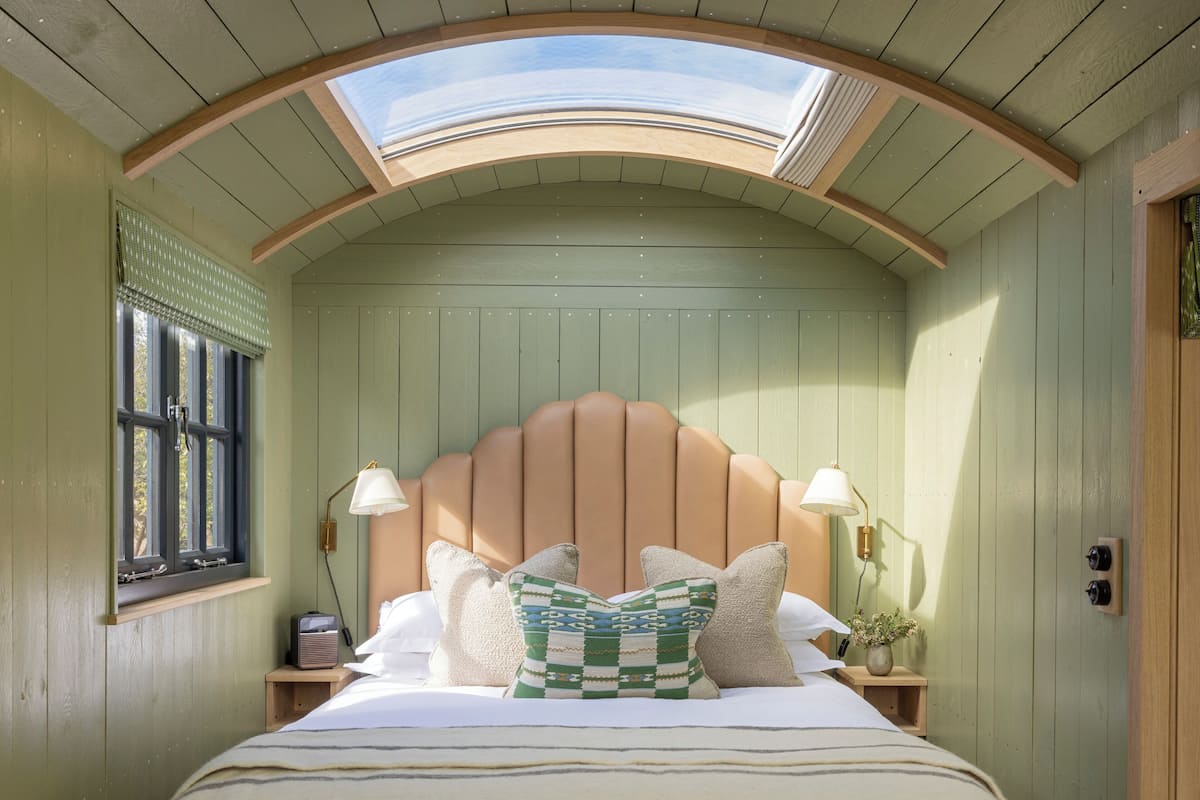 Una habitación con cama, tragaluz, ventana con persianas y mesita de noche con lámpara.
