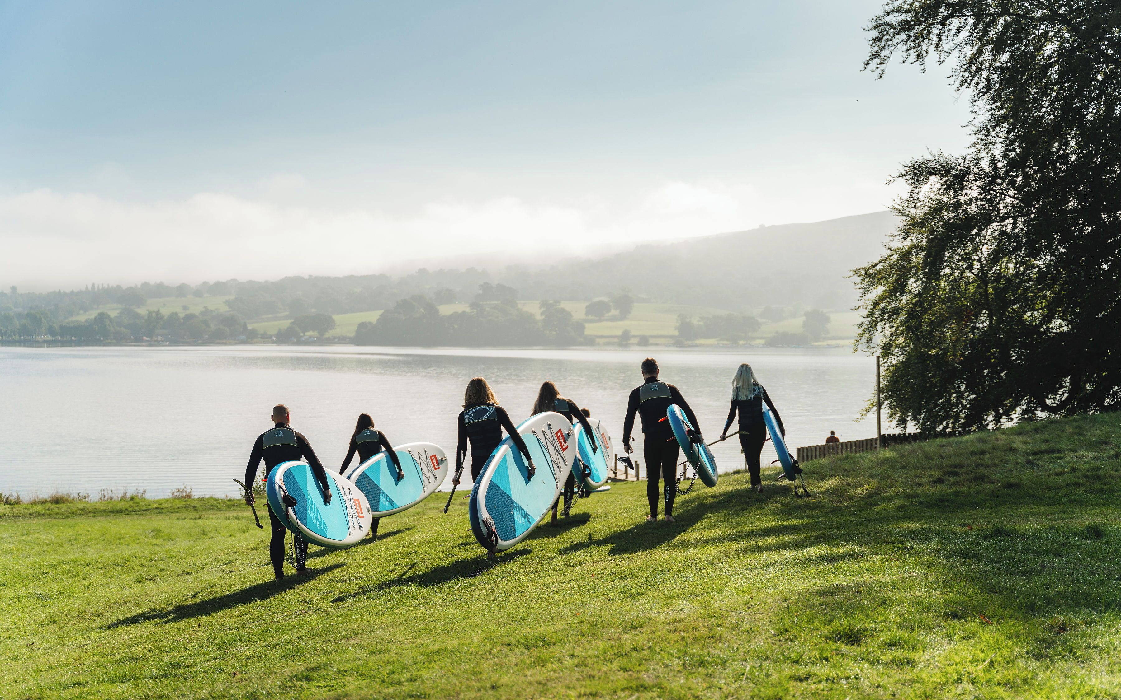 A group of people with surfboards walking on a grassy path beside a lake.