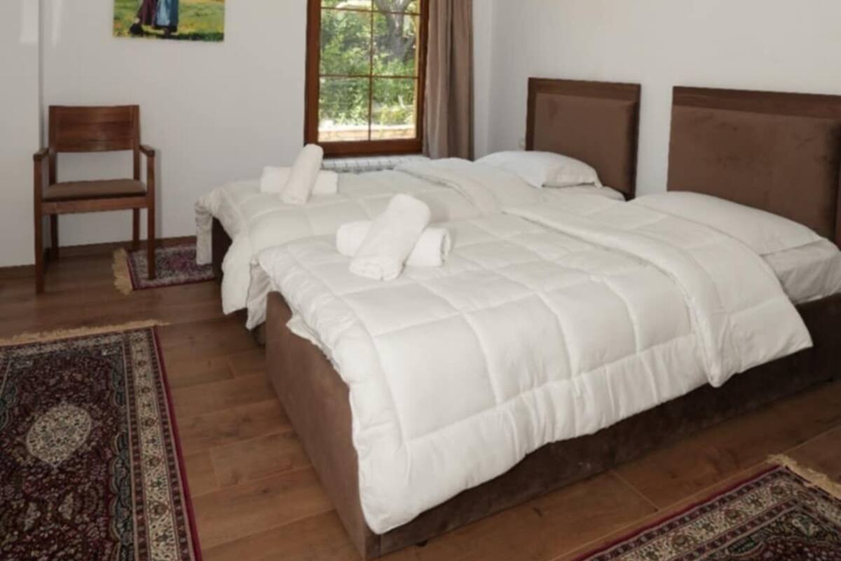 A bedroom with a large bed, a chair, a window with curtains, a painting on the wall, and a ceiling fan.