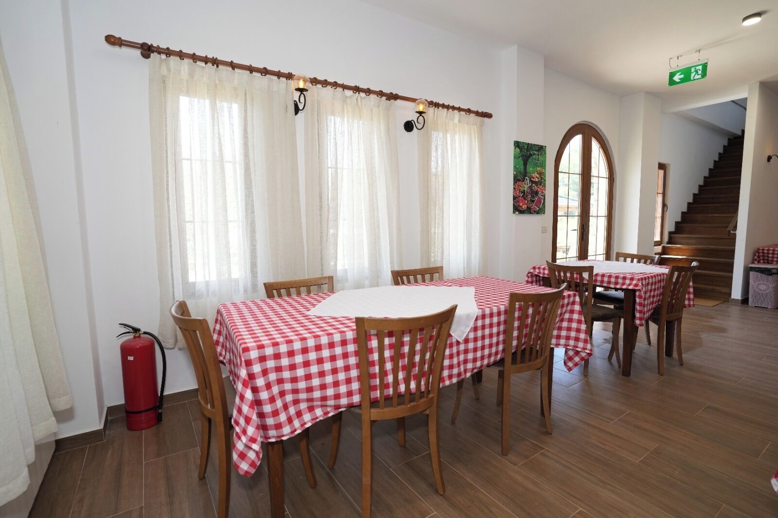 A dining area with a table set for four, wooden chairs, a fire extinguisher, and a staircase in the background.
