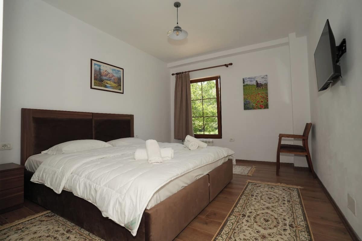A bedroom with a large bed, a wooden nightstand, a chair, a TV mounted on the wall, and a window with curtains.