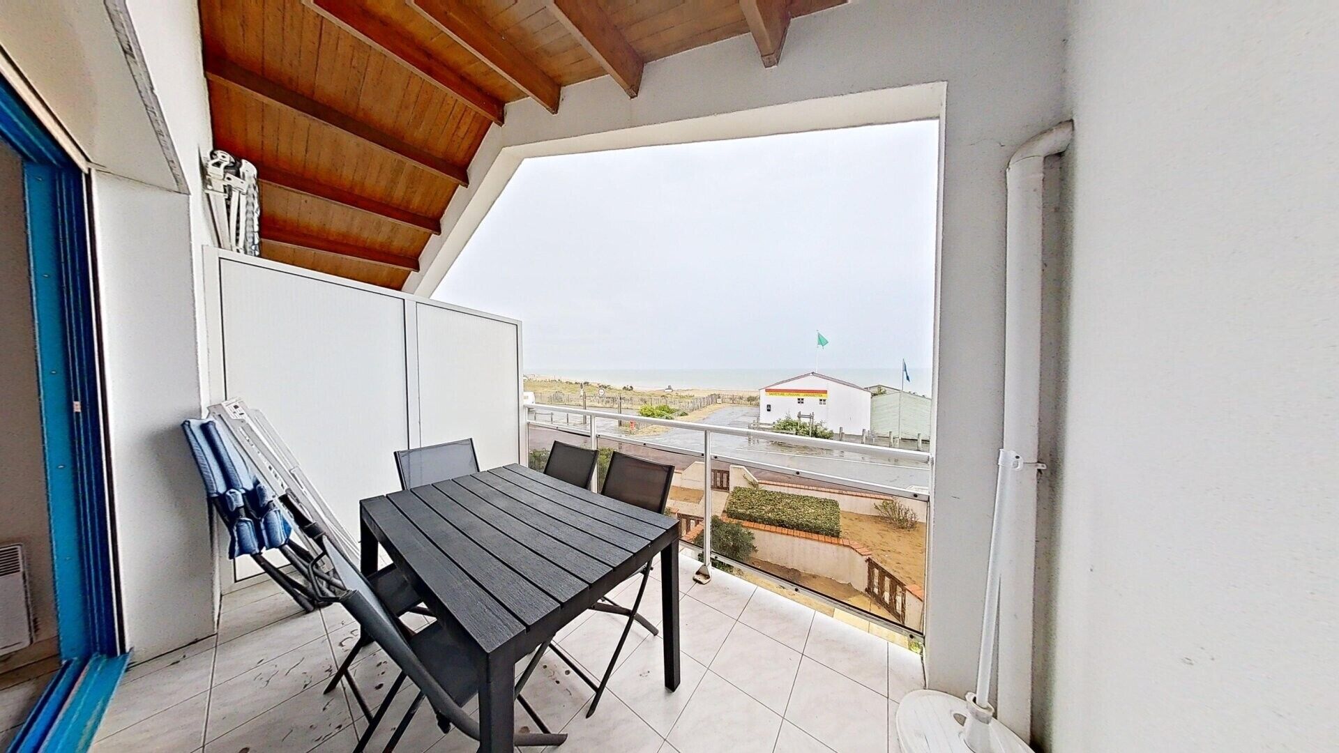 Un balcón con mesa y sillas, vista a la playa y un edificio al fondo.