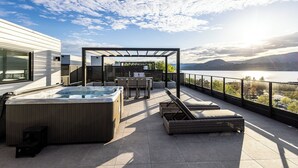 En takterrass med ett bubbelbad, solstolar och en pergola med utsikt över en sjö.