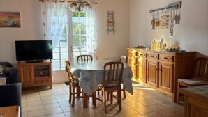 A dining area with a wooden table and chairs, a television, and a kitchen area with wooden cabinets. - Maison Malovyn: Quiet House Near Beach and Forest (La Tranche-sur-Mer)