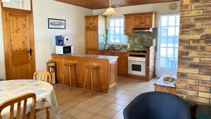 A kitchen with wooden cabinets, a microwave, a stove, and a sink. There is a dining table with chairs and a blue chair. - Maison Malovyn: Quiet House Near Beach and Forest (La Tranche-sur-Mer)