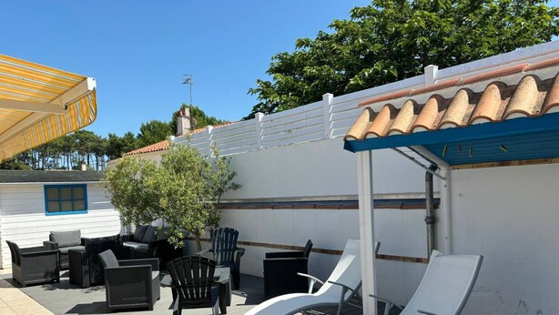 An outdoor patio with wicker furniture and white lounge chairs, a white building with blue trim, and a tiled roof. - Maison Malovyn: Quiet House Near Beach and Forest (La Tranche-sur-Mer)