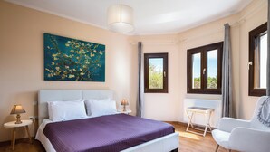 Un dormitorio con cama, una silla, una mesita, un cuadro y un ventanal.